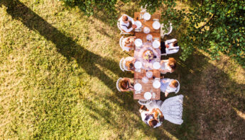 Wedding reception outside in the backyard. Bride and groom with a family around the table, kissing each other. Aerial view.